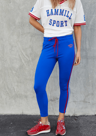 Woman wearing a white Hammill Sport jersey, blue athletic leggings, and red sneakers.