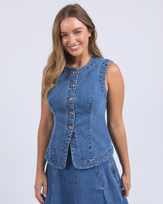 Woman wearing a sleeveless blue denim button-up vest and matching denim skirt.