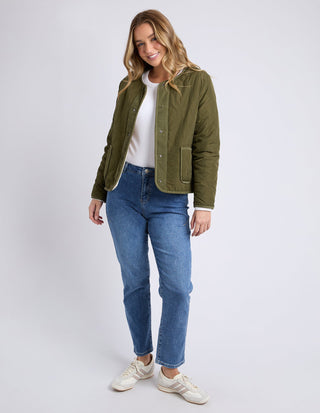 Woman wearing an olive green quilted jacket, white t-shirt, and blue denim jeans.