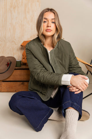Woman wearing an olive green bomber jacket and navy wide-leg pants sitting on the floor.