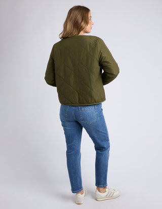 Woman wearing an olive green quilted jacket and blue denim jeans standing against a grey background.