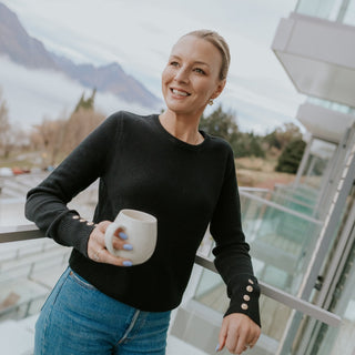 Woman in a black long-sleeve knit sweater and blue jeans holding a white mug on a balcony.