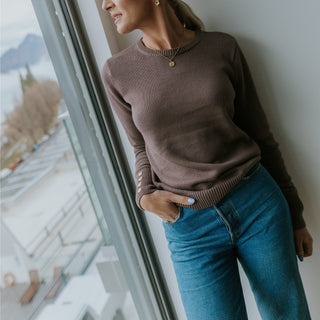Woman wearing a brown ribbed long sleeve top and blue jeans leaning against a window.