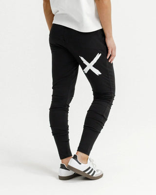Woman wearing black leisure pants with a white X graphic and white sneakers.