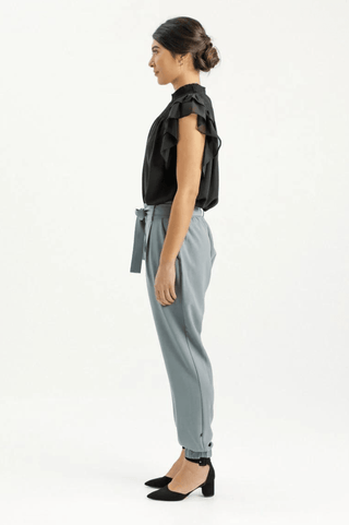 Woman in a black sleeveless ruffle blouse and grey high-waisted tapered trousers.