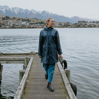 Woman wearing a navy blue waterproof raincoat and blue jeans walking on a wooden pier by a lake.
