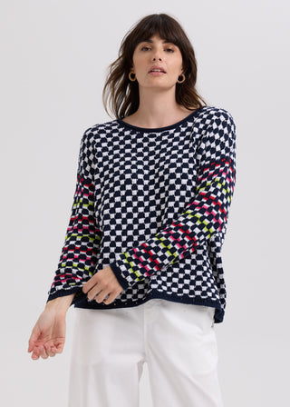 Woman wearing a navy and white checkered knit sweater with colorful striped sleeves and white pants