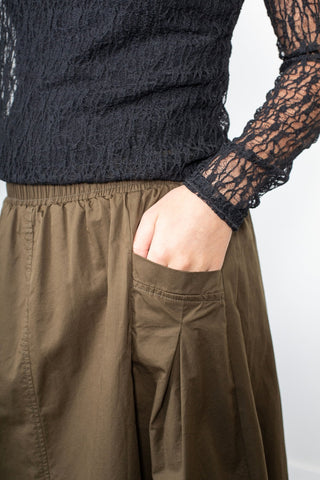 Woman wearing a black lace long sleeve top and olive green pleated skirt with pockets.