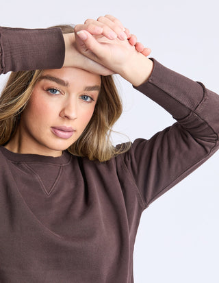 Woman wearing a brown long-sleeve sweatshirt posing with hands on her forehead.