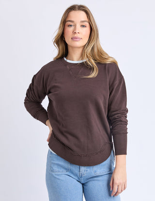 Woman wearing a brown long-sleeved sweatshirt with raw hem and blue denim jeans.