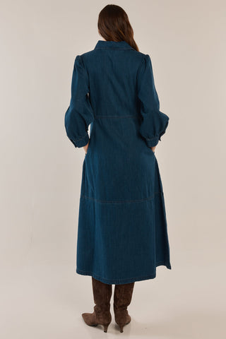 Rear view of a woman wearing a long-sleeved blue denim maxi dress and brown boots.