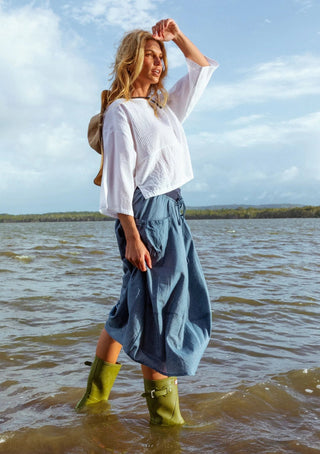 Woman wearing a white blouse, blue linen skirt, and green rain boots by the water.