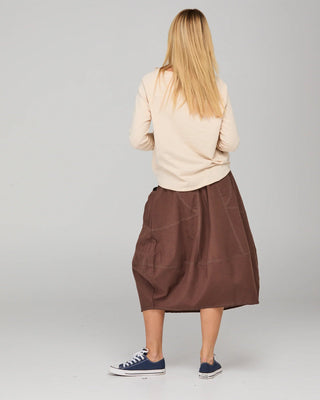Woman in a cream long-sleeve top and brown midi skirt with blue canvas sneakers.