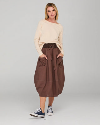 Woman wearing a cream long sleeve top and brown midi skirt with pockets and blue sneakers.