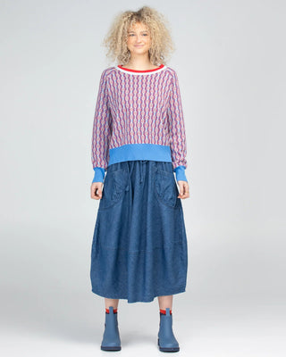 Woman wearing a patterned red and blue sweatshirt with a long denim skirt and blue boots.