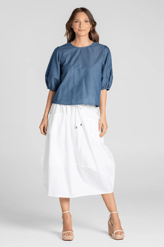 Woman wearing a blue denim puff sleeve top and white midi skirt with wedge sandals.
