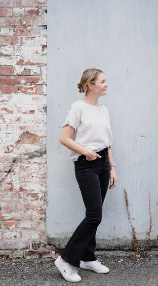 Woman wearing a white textured blouse and black flared jeans standing by a brick wall.