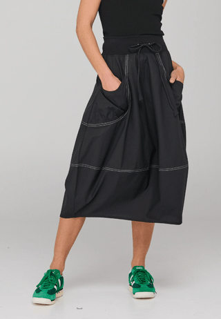 Woman wearing a black midi skirt with contrast white stitching, pockets, and green sneakers.