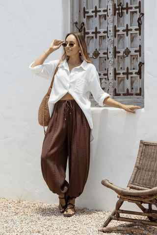 Woman wearing a white linen shirt and brown drawstring harem pants with a straw bag.