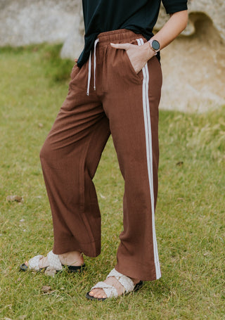 Woman wearing brown wide-leg leisure pants with white side stripes and braided sandals.