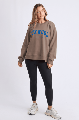 Woman wearing a brown Foxwood graphic sweatshirt, black leggings, and white sneakers.