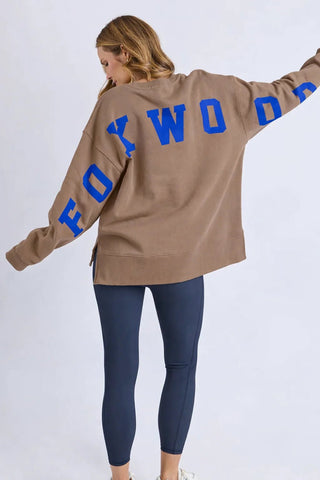 Woman wearing a tan Foxwood oversized sweatshirt with blue lettering and dark leggings.