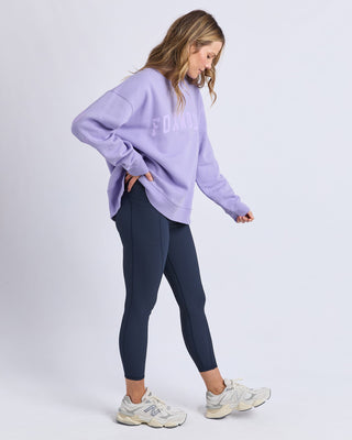 Woman wearing a lilac Foxwood sweatshirt, navy blue leggings, and white sneakers.