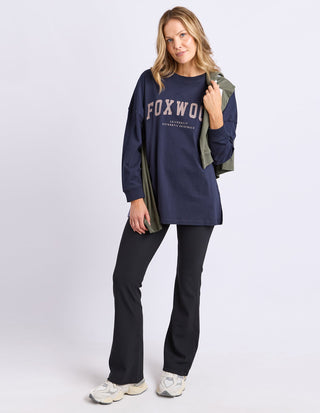 Woman wearing a navy Foxwood long sleeve sweatshirt and black flared pants.