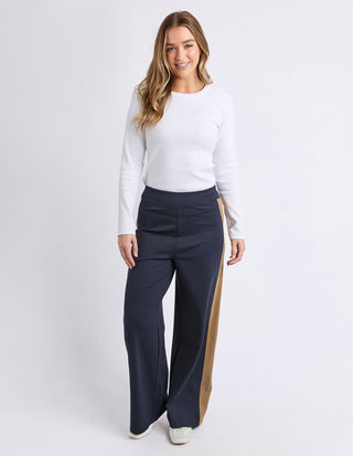 Woman wearing a white long-sleeve top and navy wide-leg trousers with tan side stripes.