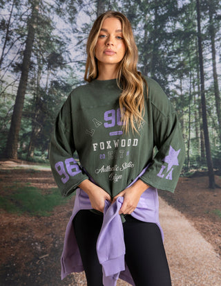 Woman wearing a green Foxwood oversized sweatshirt and purple leisure wear in a forest setting.