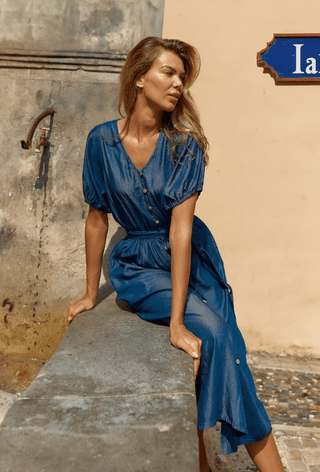 Woman wearing a blue short sleeve button-up midi dress sitting on a stone ledge outdoors.
