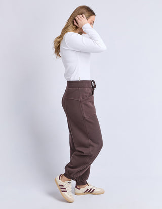 Woman wearing brown Homelee Apartment pants and a white long sleeve top with cream sneakers.