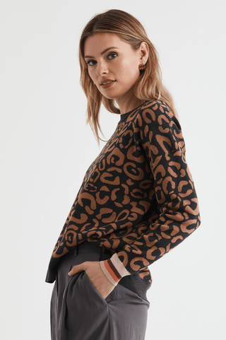 Woman wearing a black and brown leopard print sweater with striped cuffs and grey pants.