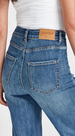 Woman wearing blue denim jeans with a brown Junkfood brand waist patch and white top.