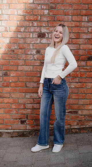 Smiling blonde woman wearing a white long-sleeve top and blue wide-leg jeans against a brick wall
