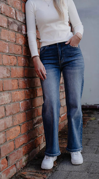 Woman wearing a white long-sleeve top and blue wide-leg denim jeans leaning against a brick wall.