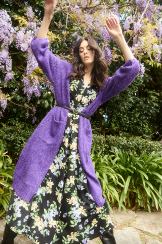 Woman wearing a purple knit coatigan over a black floral maxi dress with a black belt.