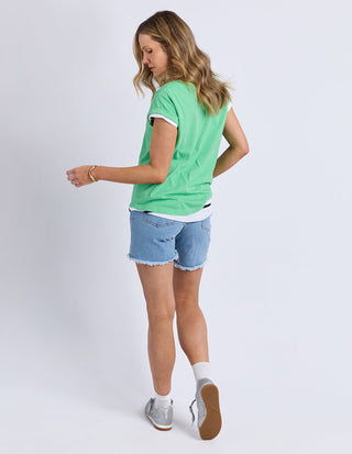 Woman wearing a green t-shirt, denim shorts, and grey sneakers standing against a white background