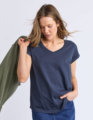 Woman wearing a navy blue v-neck t-shirt and blue jeans holding an olive green sweatshirt.