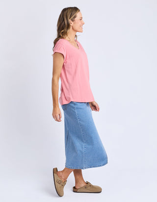 Woman wearing a pink short-sleeved V-neck top, light blue denim midi skirt, and tan clogs.