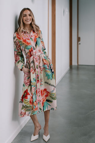 Woman wearing a pink floral print midi shirt dress with a waist tie and white heels.