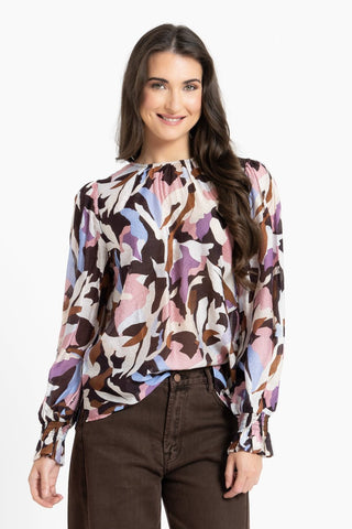 Woman wearing a multicolored abstract print long sleeve blouse and brown trousers.