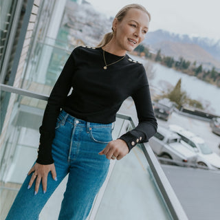 Woman wearing a black long-sleeve top with gold button details and blue denim jeans.