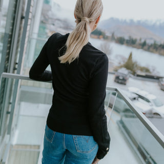 Woman in a black long-sleeve top and blue jeans standing on a balcony overlooking a lake.