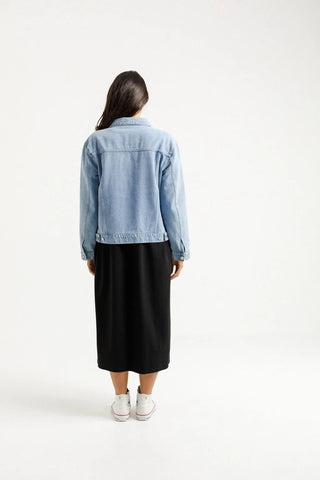 Back view of a woman wearing a light blue denim jacket and a black midi skirt.