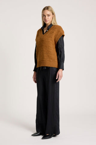 Woman wearing a brown cable knit vest over a black button-down shirt and wide-leg trousers.