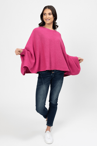 Woman wearing a bright pink oversized knit poncho with dark blue jeans and white sneakers.