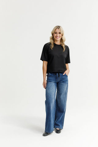 Woman smiling in a black short sleeve t-shirt and blue wide leg denim pants.