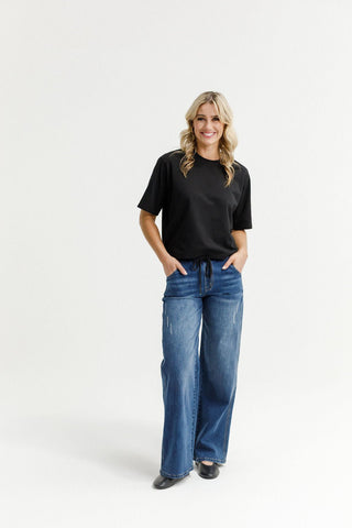Woman wearing a black t-shirt and blue wide-leg denim pants standing against a white background.