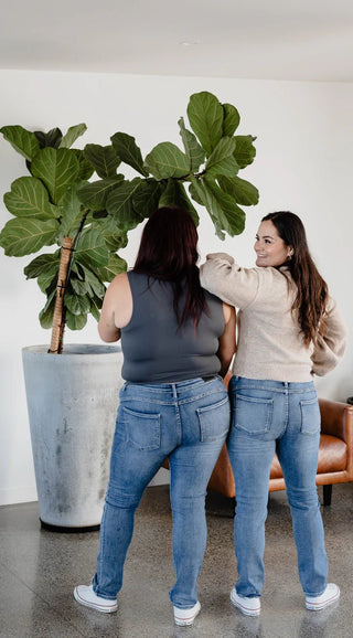 Two women wearing casual blue jeans and a grey tank top and beige sweater standing by a plant.